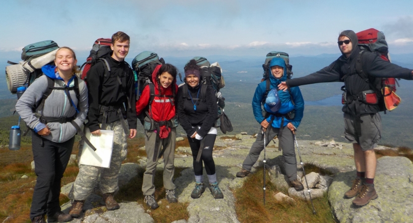 A group of students wearing backpacks stand on a rocky overlook high above the terrain below. 